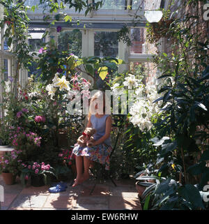 Kleine Mädchen sitzen in einem Wintergarten voller blühender Pflanzen Stockfoto