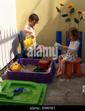 Zwei kleine Mädchen sitzen auf Stühlen und spielen neben einem Kunststoff Sandkasten im Garten Stockfoto