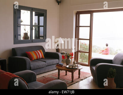 Grauen Sofa unter internen Fenster im marokkanischen Wohnzimmer mit Blick durch Glastüren Frau sitzt auf Hof Stockfoto