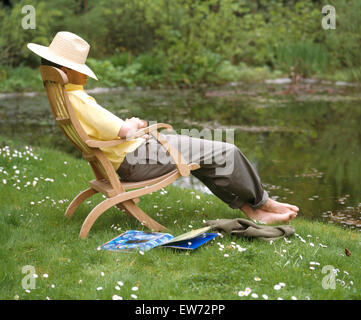 Junge Mann trägt einen Strohhut sitzen und auf einem hölzernen Liegestuhl dösen Stockfoto