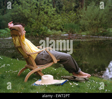 Junger Mann sitzen und dösen auf einer Chaiselongue neben einem Gartenpool Stockfoto