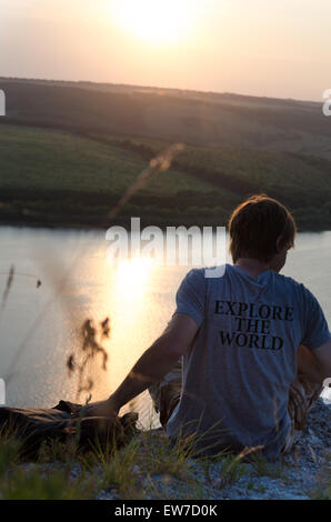 Mann sitzt auf einem Felsen über dem Fluss bei Sonnenuntergang, Reise-Konzept. Entdecken Sie die Welt - auf dem T-shirt geschrieben Stockfoto