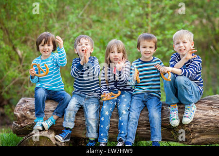 Fünf bezaubernde Kinder, gekleidet in gestreiften Hemden, auf hölzernen Baumstamm sitzend Essen Brezeln für Nachmittags-Snack, lächelnd und ch Stockfoto