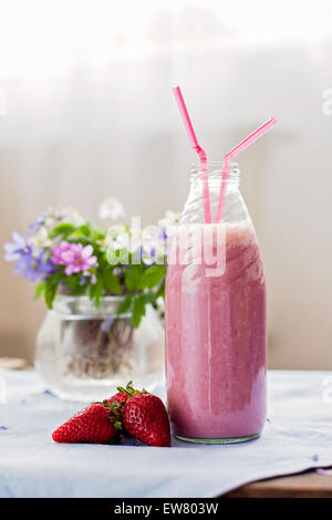 Erdbeer Smoothie frisch zubereitet in einem Glas mit einer gefütterten Stroh, Wald Frühlingsblumen in einer Vase hinter der Flasche Stockfoto