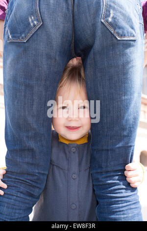 Blondine neunzehn Monate Alter Baby mit blauen und gelben Kleid zwischen Mutter Blue Jeans zurück Frau Beine Blick in die Kamera Stockfoto