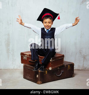 kleinen schwarzen Jungen in Graduation Hut sitzt auf dem alten Koffer und Schaukel Hände Stockfoto