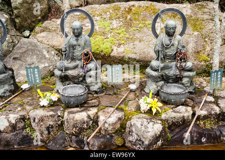 Japan, Miyajima. Daisho-in-Tempel. Zwei Jizo Bosatsu-Steinstatuen buddhistischer Mönch, die Zepter halten, Shakujo. Vordergrund, drei Pfannen auf Felsen. Stockfoto