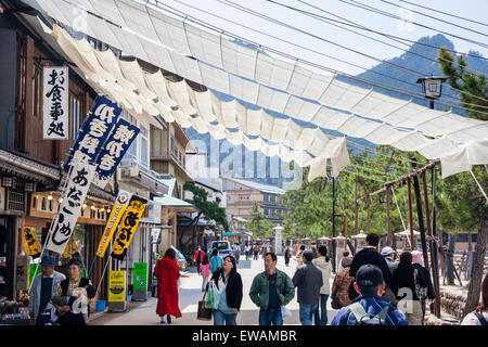 Touristen gehen unter Sonnenschirmen über eine Straße mit Souvenierläden an die touristische Attraktion der Insel Miyajima in Japan gefüllt gezogen. Stockfoto