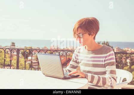 Frau mit Brille und lässige Kleidung arbeiten am Laptop im Freien auf der Terrasse. Konzept von zu Hause aus arbeiten. Natürliches Tageslicht. Stockfoto