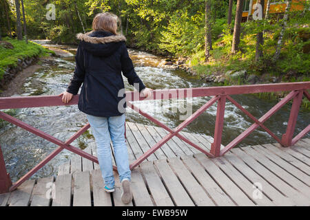 Schöne blonde kaukasische Teenager-Mädchen steht auf Holzbrücke über kleine schnelle Fluss in Finnland, Rückansicht Stockfoto