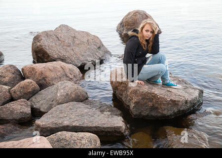 Schöne blonde kaukasische Teenager-Mädchen sitzt auf küstennahen Steinen entlang der See in Finnland Stockfoto