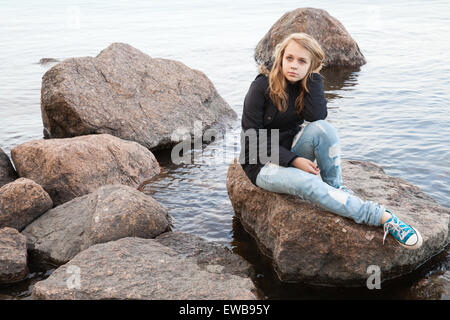 Schöne blonde kaukasische Teenager-Mädchen sitzen auf küstennahen Steinen entlang der See in Finnland Stockfoto