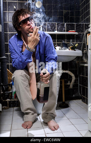 Betrunkener Mann sitzt in einer Toilette mit einer Flasche Whisky und Rauchen, Farbbild Stockfoto