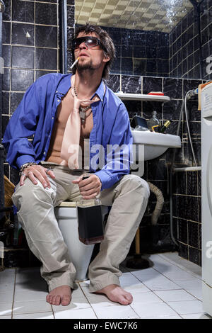 Betrunkener Mann sitzt in einer Toilette mit einer Flasche Whisky, Farbbild Stockfoto