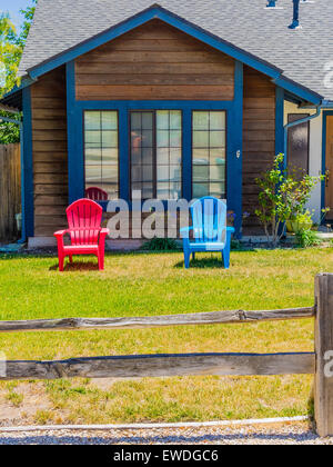 Zwei bunten Liegestühlen, einem roten und das andere blau, stehen auf dem Rasen eines Holzhauses. Stockfoto