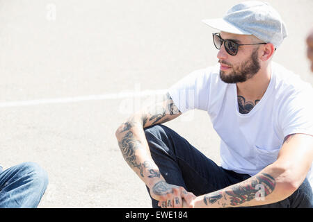 Junger Mann mit Tattoos, trägt eine Kappe und Sonnenbrille, sitzen auf dem Boden Stockfoto