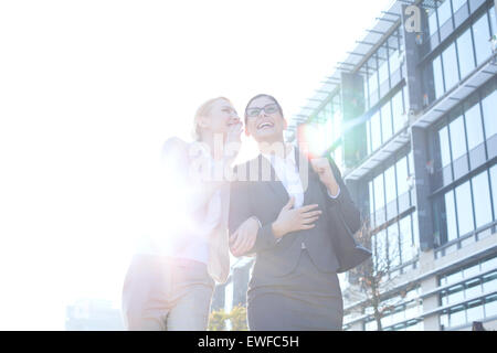 Glücklich Geschäftsfrau Kollegen Ohr außerhalb Bürogebäude am sonnigen Tag zu flüstern Stockfoto