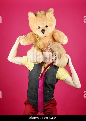 Expressive Hipster trägt eine Weste und rosa Krawatte und hält er Teddy-Bär, rosa Hintergrund und Studio gedreht Stockfoto