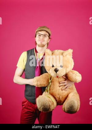Expressive Hipster trägt eine Weste und rosa Krawatte und hält er Teddy-Bär, rosa Hintergrund und Studio gedreht Stockfoto