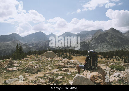 Ein Wanderer mit Rucksack sitzt auf einem großen Felsen und bestaunt die weitläufige Bergkette in der Ferne. Die Szene vermittelt ein Gefühl von Ruhe und Abenteuer in der freien Natur. Stockfoto