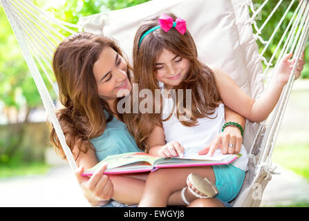 Portrait der schönen Mutter mit niedlichen kleinen Tochter sitzen auf Hinterhof in Hängematte und lesen lustige Geschichte, glückliches Familienleben Stockfoto