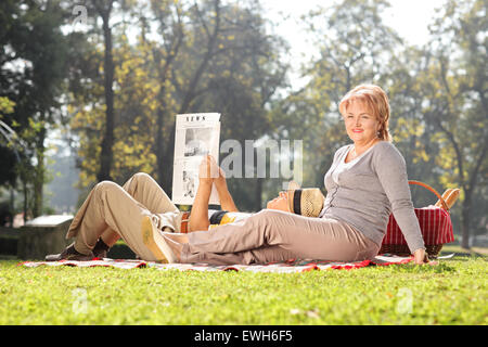 Reife Frau posiert in einem Park mit ihrem Mann auf dem Schoß liegen und liest eine Zeitung an einem schönen sonnigen Tag Stockfoto