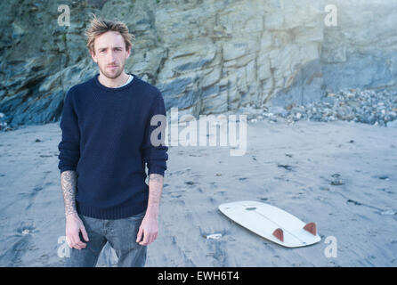 Porträt einer Surfer am Strand mit seinem Surfbrett auf dem Sand mit Sonnenlicht Flare auf den Felsen. Stockfoto