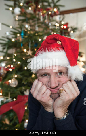 Ein mürrischer Mann in eine Weihnachtsmütze vor dem Weihnachtsbaum Stockfoto