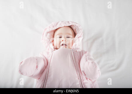 sechs Monate Alter niedlichen Baby gekleidet in rosa flauschigen Schneeanzug eng anliegende Kapuze Winterkleidung auf weißen Laken Bett lächelt glücklich fa Stockfoto