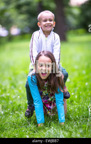 Mädchen mit ihren kleinen Bruder spielen in dem grünen Rasen. Kleiner Junge reitet auf zurück eine Schwester. Stockfoto