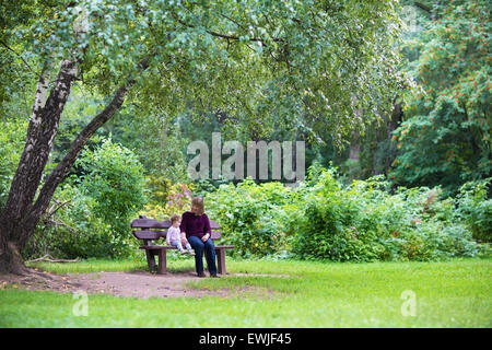 Großmutter und Baby Mädchen entspannend in einem Park auf einer Bank unter einem großen schönen Baum an einem warmen Tag im Herbst Stockfoto