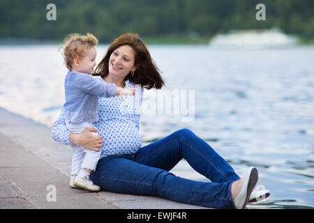 Junge schwangere Mutter und ihre kleine Baby-Tochter am Ufer eines Flusses in einem Stadtzentrum entspannend