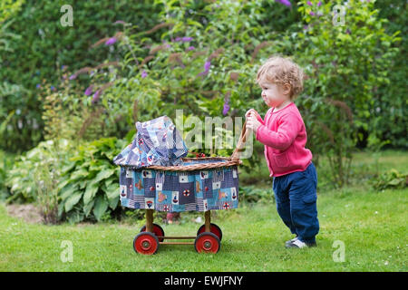 Lustige lockiges Babymädchen spielen mit einem Vintage Puppe Kinderwagen im Garten Stockfoto