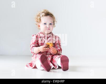 Lustige Babymädchen mit dem lockigen Haar trägt ein rotes Kleid ein Weihnachtsplätzchen in einem weißen Kindergarten Essen Stockfoto