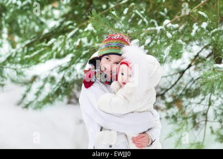 Süsser Boy umarmt seine kleine Schwester in einem schönen Winter-Park unter einem Baum im Schnee beide tragen warme weiße Jacken und c Stockfoto