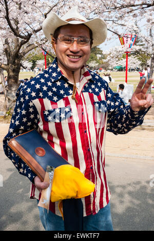 Lächelnd Japaner unter Kirschblüten in Japan posieren. Trägt Sterne und Streifen Langarm Shirt und Cowboyhut, gibt mit zwei Fingern Frieden Geste. Stockfoto