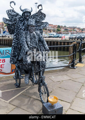 Ein Mann posiert als eine sehr aufwendige lebende Statue auf einem Dreirad und mit einem Teufel auf dem Rücken in Whitby North Yorkshire England UK Stockfoto