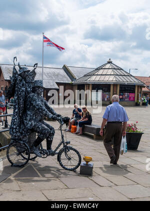 Ein Mann posiert als eine sehr aufwendige lebende Statue auf einem Dreirad und mit einem Teufel auf dem Rücken in Whitby North Yorkshire England UK Stockfoto