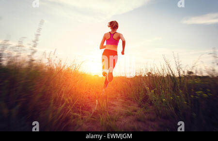 Junge Frau läuft auf einer Landstraße bei Sonnenuntergang in Sommerwiese. Lifestyle-Sport-Hintergrund Stockfoto