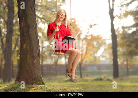 Fröhliche Mädchen in einem roten Kleid, einem Buch sitzend auf Schaukel in einem park Stockfoto