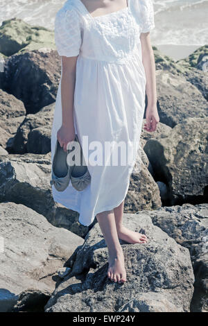 eine Mädchen steht barfuß auf einem Felsen am Meer, mit ihren Schuhen in der hand Stockfoto