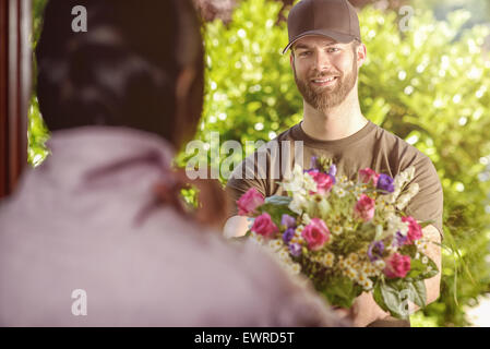Lächelnde bärtigen 20er Jahre Mann tragen braune Haube und braunen T-shirt liefert Blumen an Tür der weibliche junge Brünette. Über die Schulter Stockfoto
