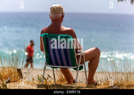 Senior Mann in einem Klappstuhl, der das Meer beobachtet. Der Strand von Plakias, Südkreta, Griechenland. Sonnenbaden Senior am Strand Sonne Wohlbefinden draußen Stockfoto