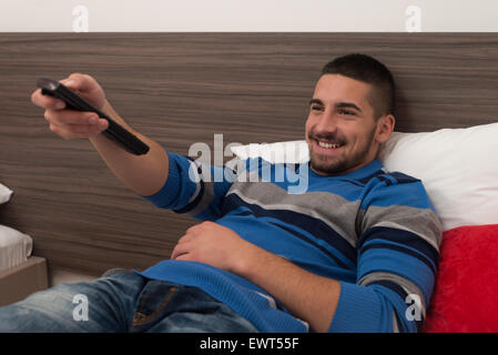 Hübscher junger Mann Fernsehen auf Bett mit der Fernbedienung In der Hand liegend Stockfoto