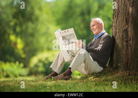 Fröhlichen senior Gentleman Zeitunglesen im Park und lehnte sich gegen einen Baum auf dem Boden sitzend an schönen Sommertag Stockfoto