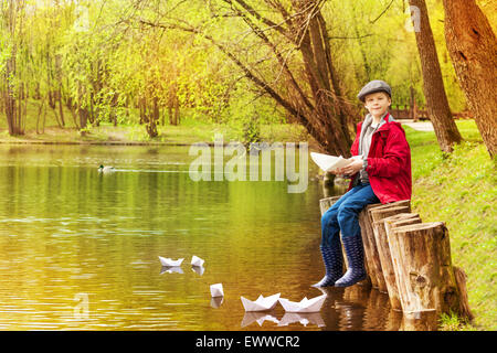 Junge sitzen in der Nähe Teich spielen mit weißem Papier Boote Stockfoto