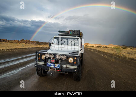 Keetmanshoop, Namibia, Afrika - Land Rover fahren auf nasser Fahrbahn mit überhängenden Regenbogen am Himmel dahinter Stockfoto