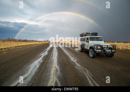 Keetmanshoop, Namibia, Afrika - Land Rover fahren auf nasser Fahrbahn mit überhängenden Regenbogen am Himmel dahinter Stockfoto