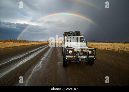 Keetmanshoop, Namibia, Afrika - Land Rover fahren auf nasser Fahrbahn mit überhängenden Regenbogen am Himmel dahinter Stockfoto