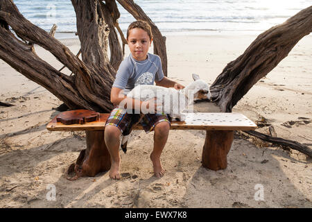 Ein junger Mann sitzt auf einem handgefertigten Holzbank mit einem Lamm auf seinem Schoß am Strand in Guanacaste Costa Rica Stockfoto